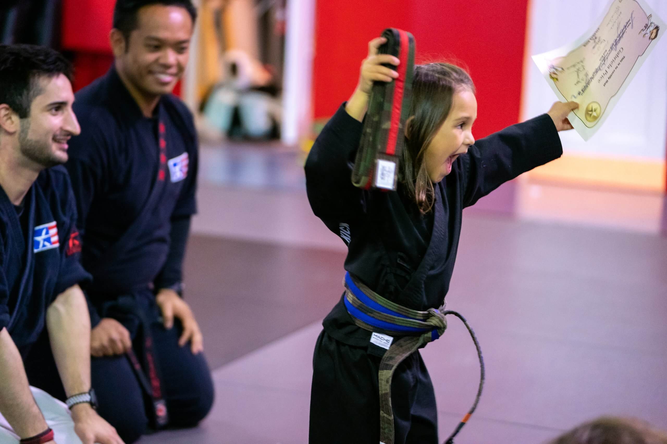 A girl in a black gi holds up a new red belt and a certificate while two instructors kneel beside her.