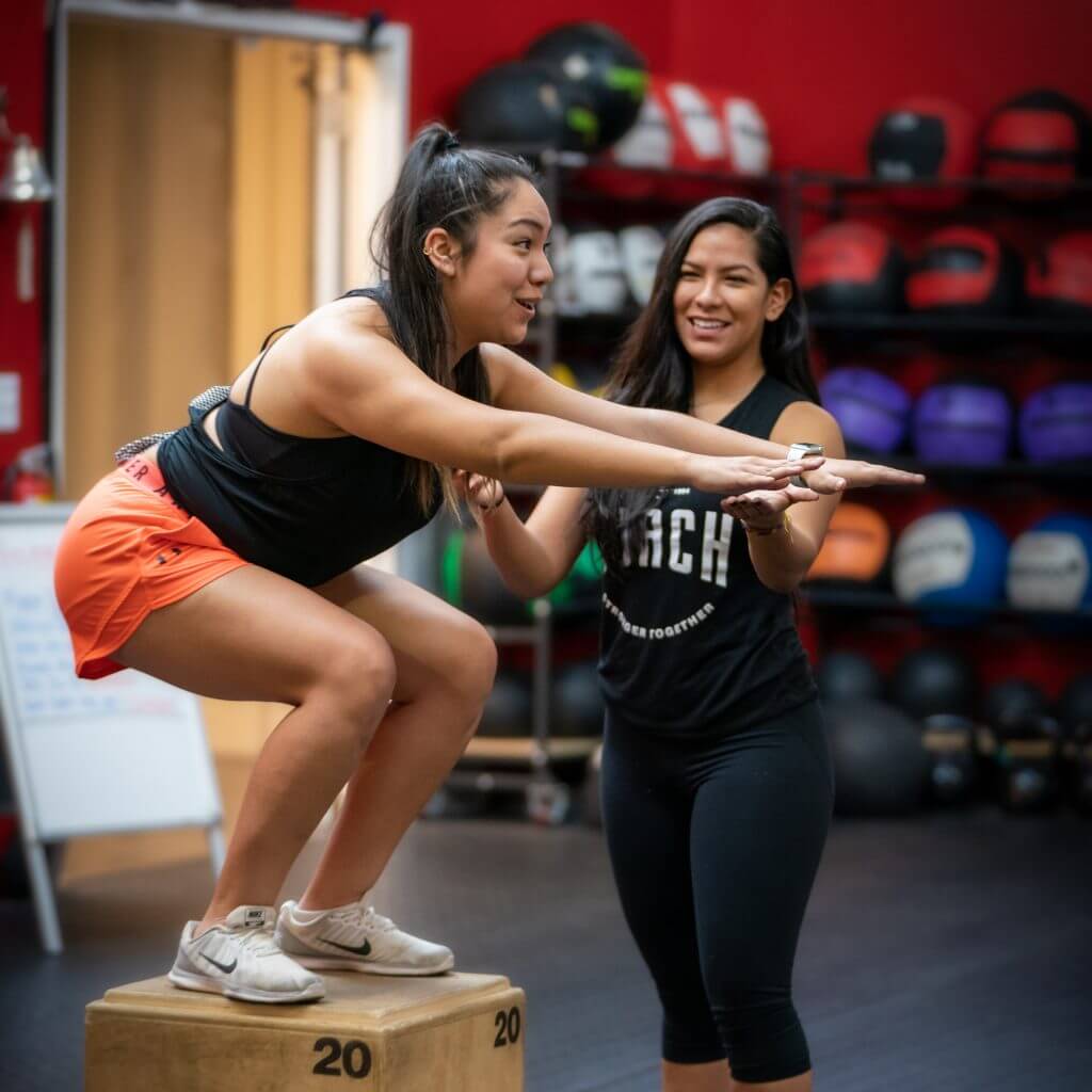 A coach spots a member balancing on a plyo box. Both faces are in frame, in front of a red wall.