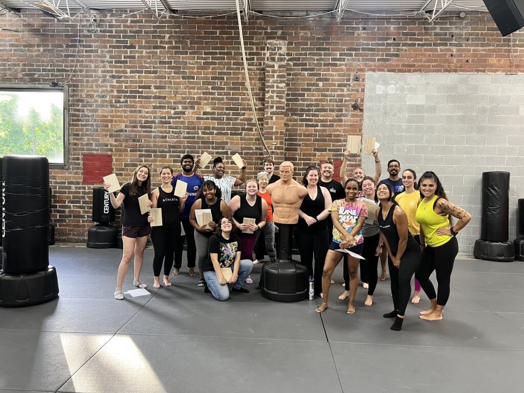 A self-defense class poses with certificates beside a training dummy. Faces and feet are in frame.