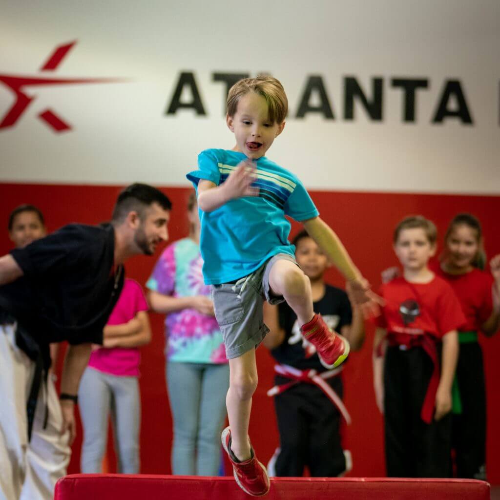 A boy in a blue shirt jumps onto a red mat during a kids class, instructors and students watching.