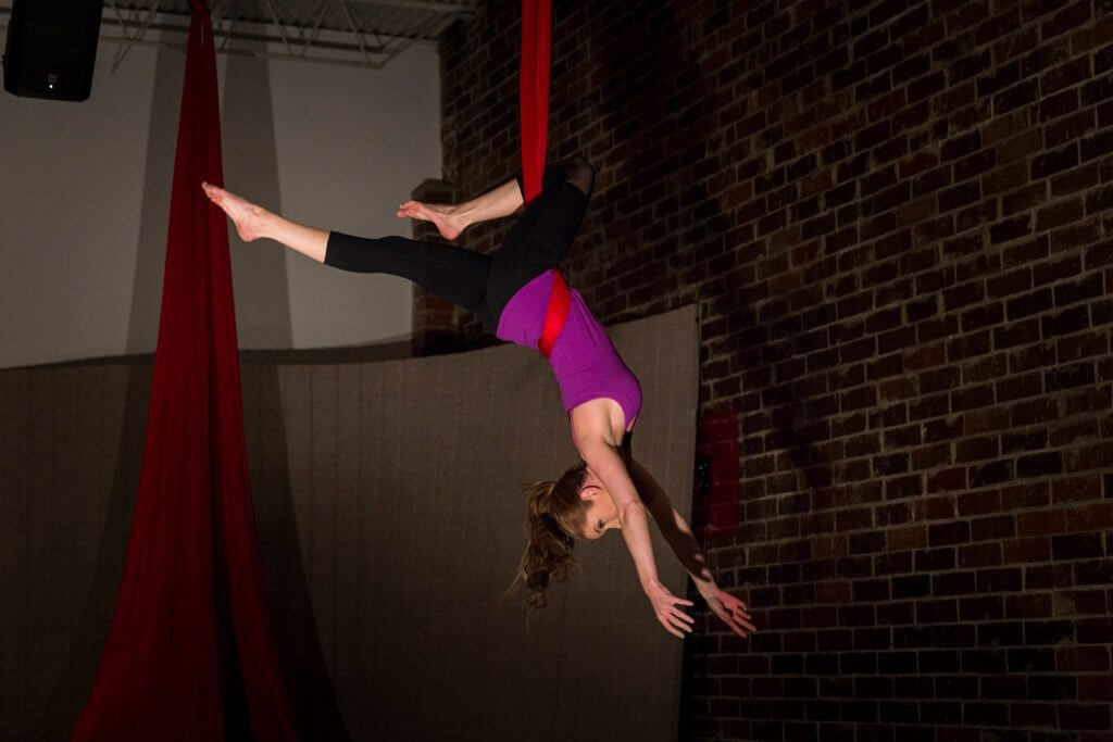 An aerialist hangs inverted in red silks, head and feet both in the frame, against a brick wall.